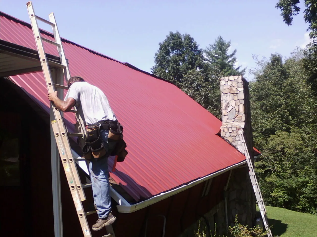 Roofer on a ladder beside a red metal roof and stone chimney — Soffit & Fascia Repair by Roofing Brothers in Richville